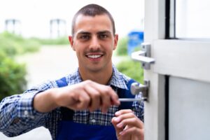 Portrait of a smiling male locksmith in a plaid shirt and blue overalls using a screwdriver to work on a commercial door lock in an outdoor setting.