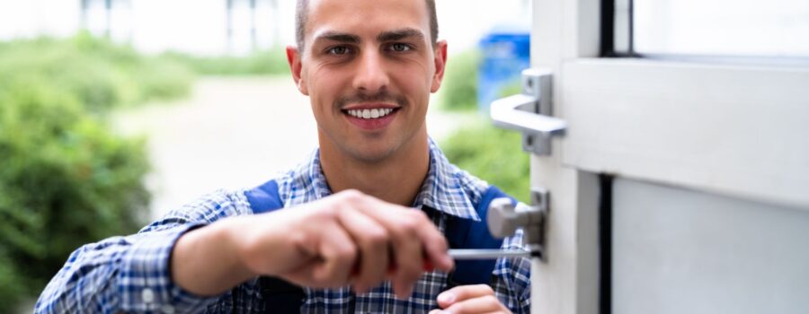 Portrait of a smiling male locksmith in a plaid shirt and blue overalls using a screwdriver to work on a commercial door lock in an outdoor setting.